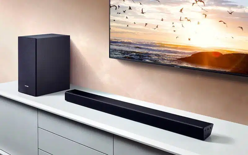 تصویری از یک ساندبار سیاه رنگ که یک سیستم صوتی می باشد و در جلوی تلویزیون قرار دارد را مشاهده می نمایید.